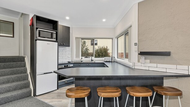 A modern kitchen with a large island, bar stools, and a view of the outdoors. - Parkwood 5 - 2BR Split Level l Spa Bath l BBQ l Central Location l WiFi (Jindabyne)