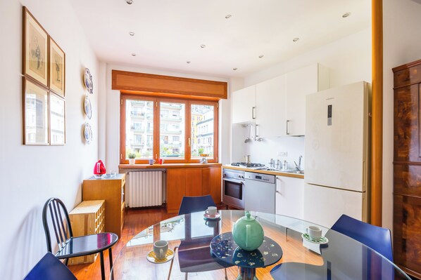 A modern kitchen with a dining area, featuring a round glass table, chairs, and a view of the city through large windows. - Colonna Classic Style Apartment by Wonderful Italy (Naples)