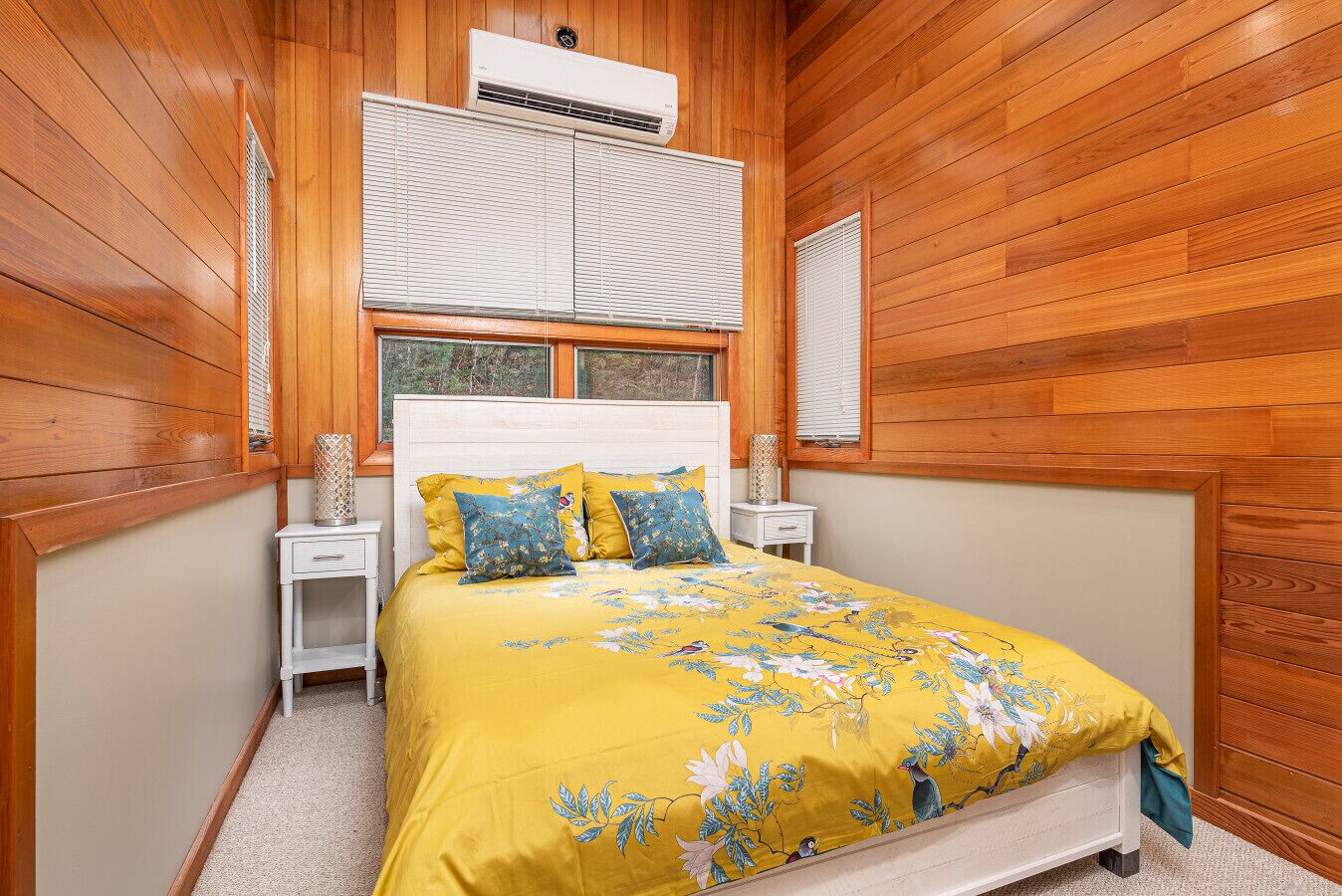 Une chambre avec des murs en bois, un lit recouvert d’une courtepointe à motifs floraux jaunes, deux tables de chevet et un appareil de climatisation placé au-dessus de la fenêtre.