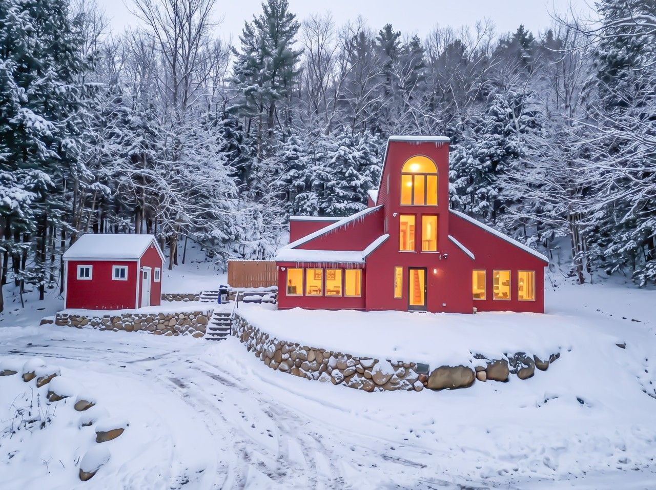 A red building with a stone wall and a smaller red structure nearby, surrounded by snow-covered trees.