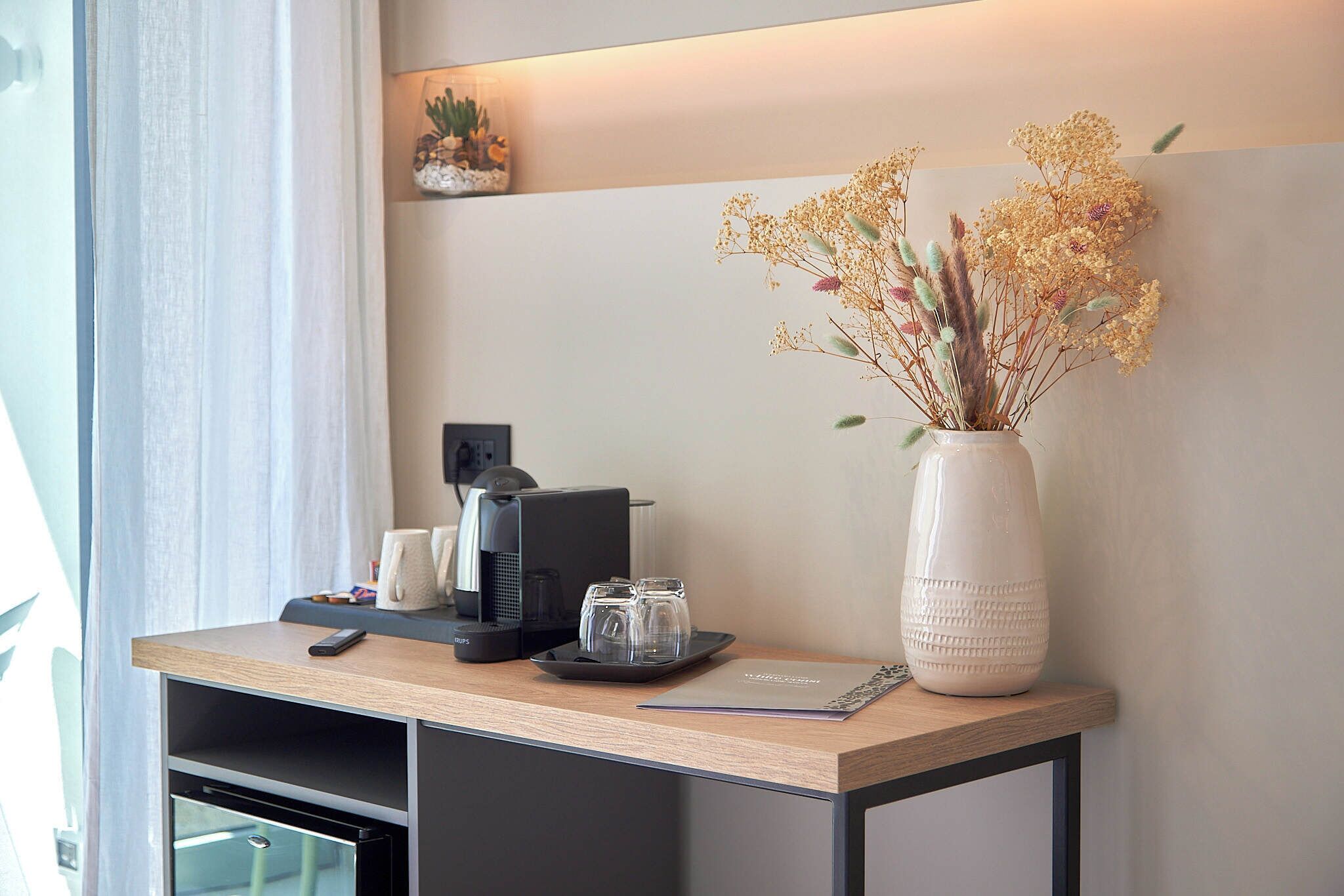 A hotel reception desk with a coffee machine, a vase with dried flowers, and a small cactus plant on a shelf.