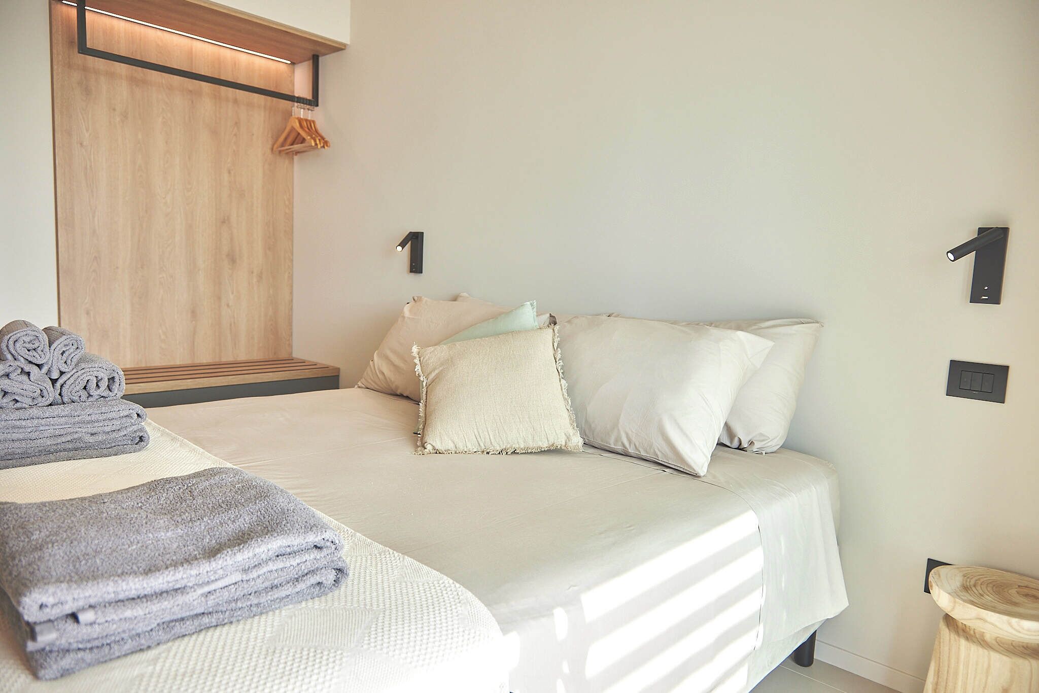 Una cama bien hecha con almohadas y una toalla doblada, un cabecero de madera y un taburete pequeño de madera.
