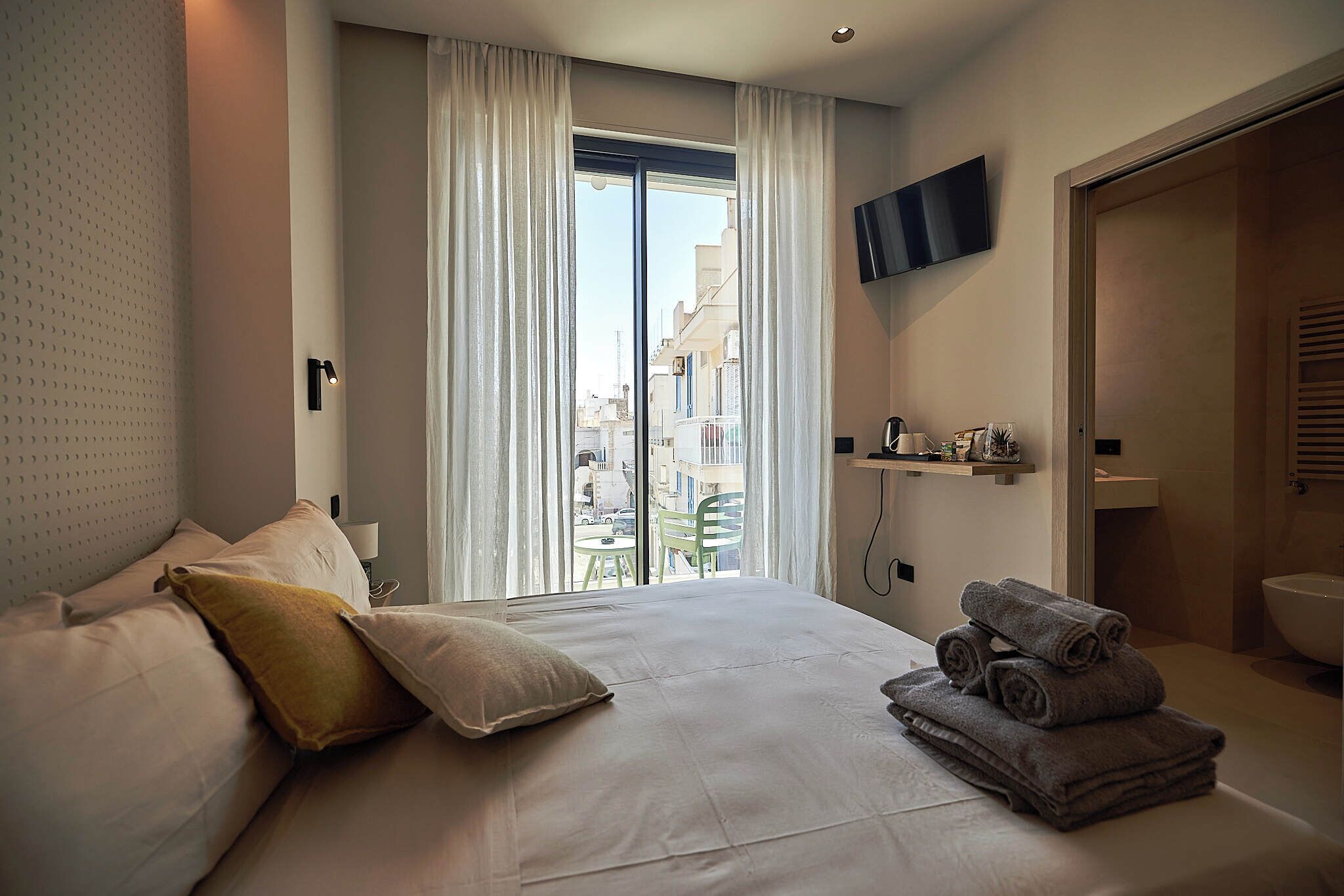 A hotel room with a bed, a TV mounted on the wall, a window with curtains, and a view of the city.