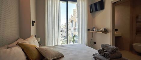 A hotel room with a bed, a TV mounted on the wall, a window with curtains, and a view of the city.