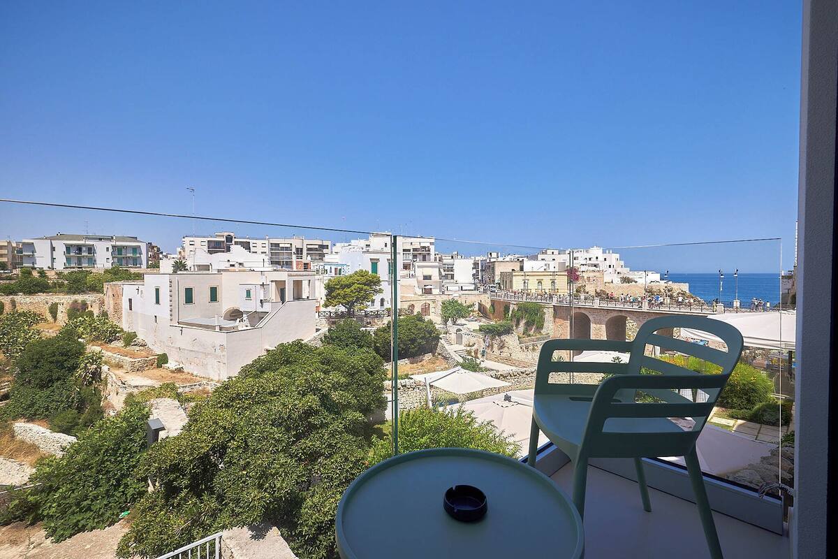 A balcony with a table and chairs overlooking a coastal town with white buildings and a clear blue sky.