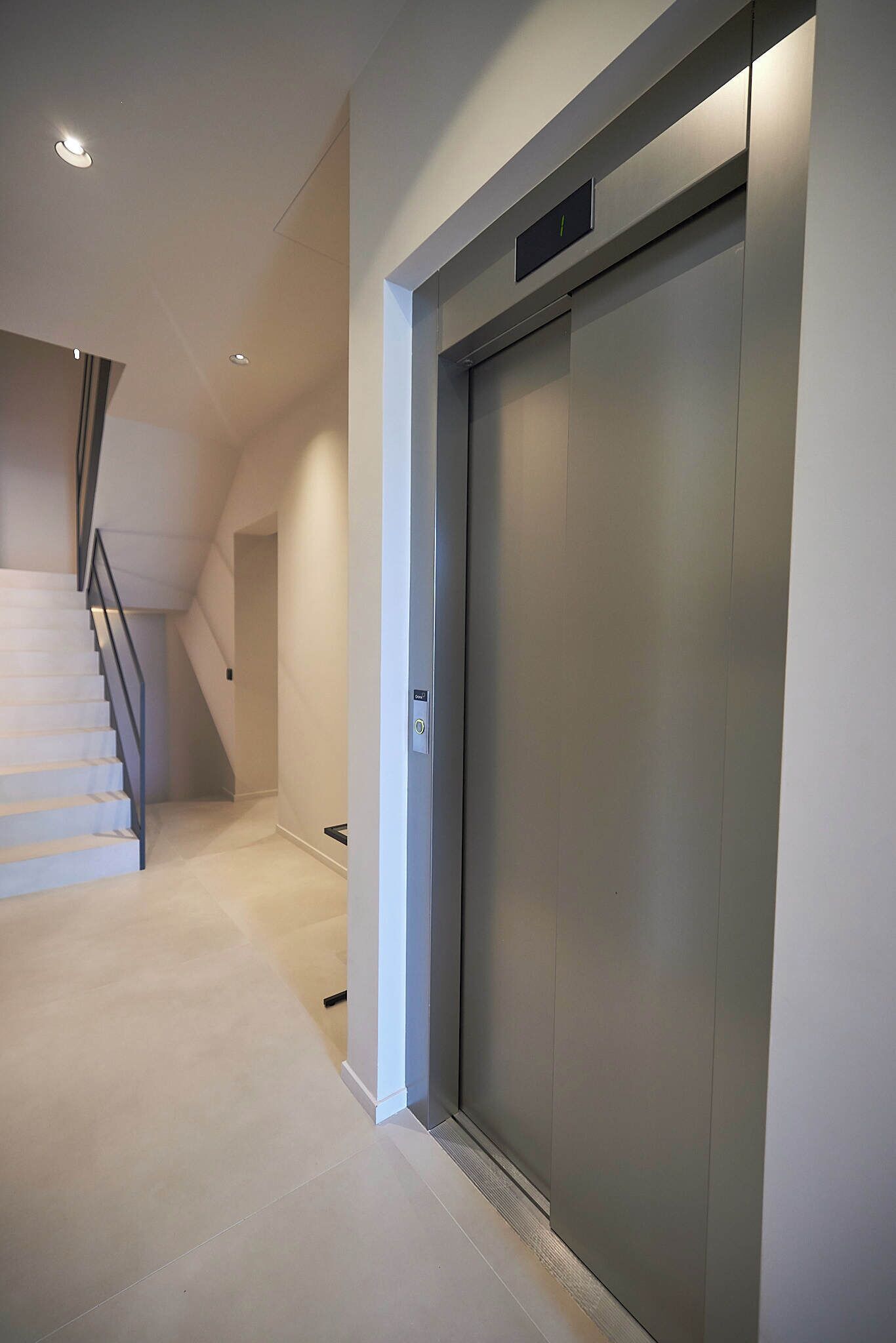 A modern elevator with a metallic finish, a staircase with a metal railing, and a clean, light-colored floor.