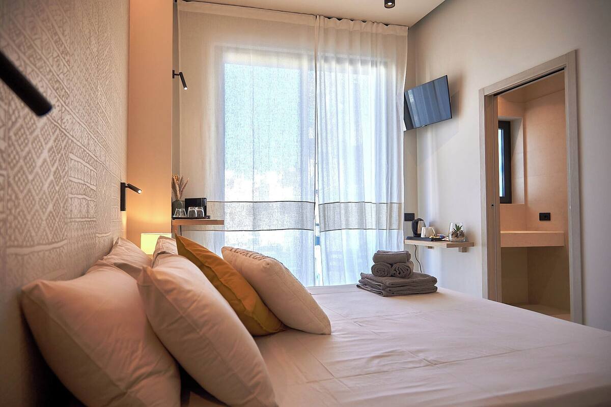 A modern bedroom with a neatly made bed, a television mounted on the wall, and a window with sheer curtains.