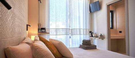 A modern bedroom with a neatly made bed, a television mounted on the wall, and a window with sheer curtains.