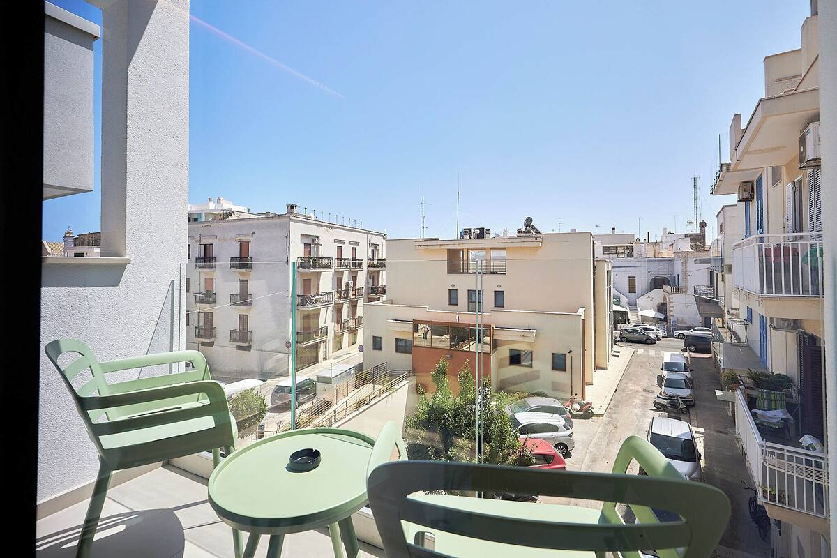 A balcony with green chairs and a table, overlooking a street with parked cars and buildings.