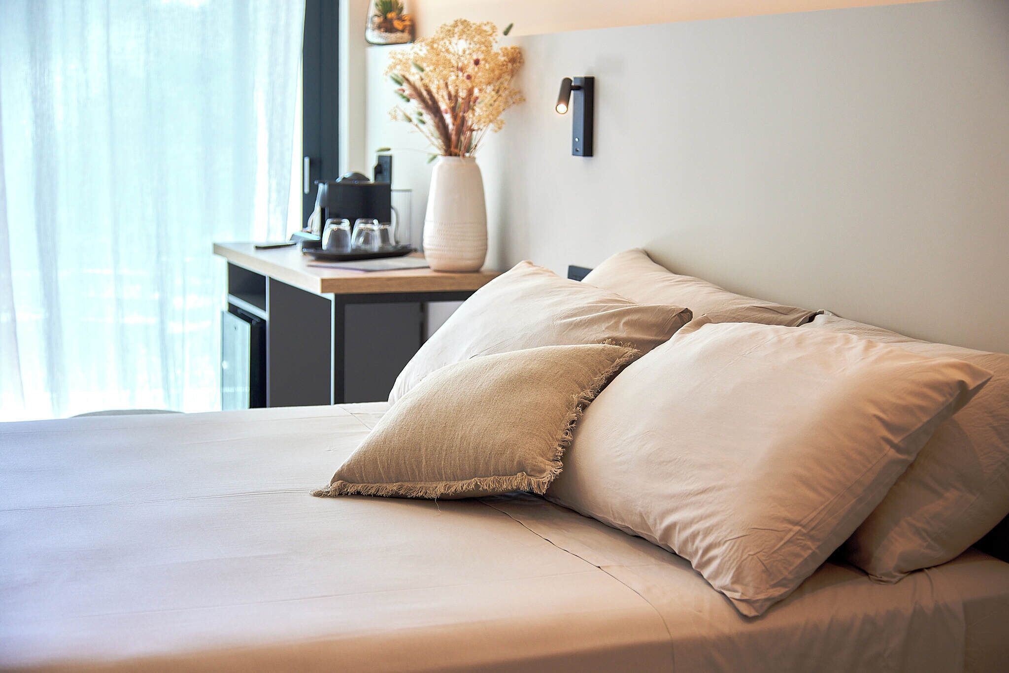 A neatly made bed with two pillows, a bedside table with a vase of dried flowers, and a wall-mounted light fixture.