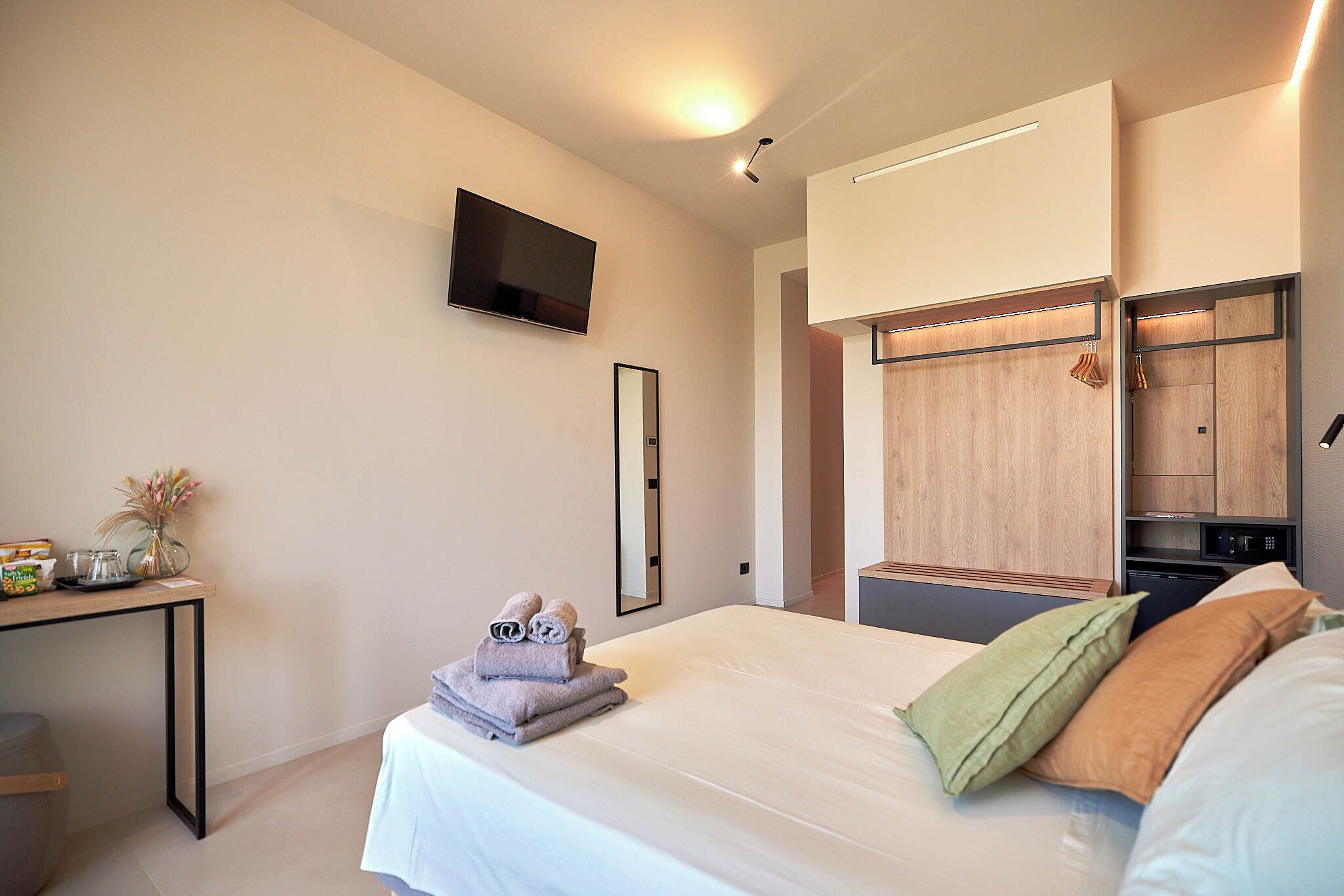 Ein modernes Hotelzimmer mit einem Bett, einem an der Wand befestigten Fernseher, einem Holzschrank und einem kleinen Tisch mit Handtüchern und einer Vase.