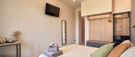 Quarto de hotel moderno com cama, televisão presa à parede, guarda-roupa de madeira e uma pequena mesa com toalhas e um vaso.