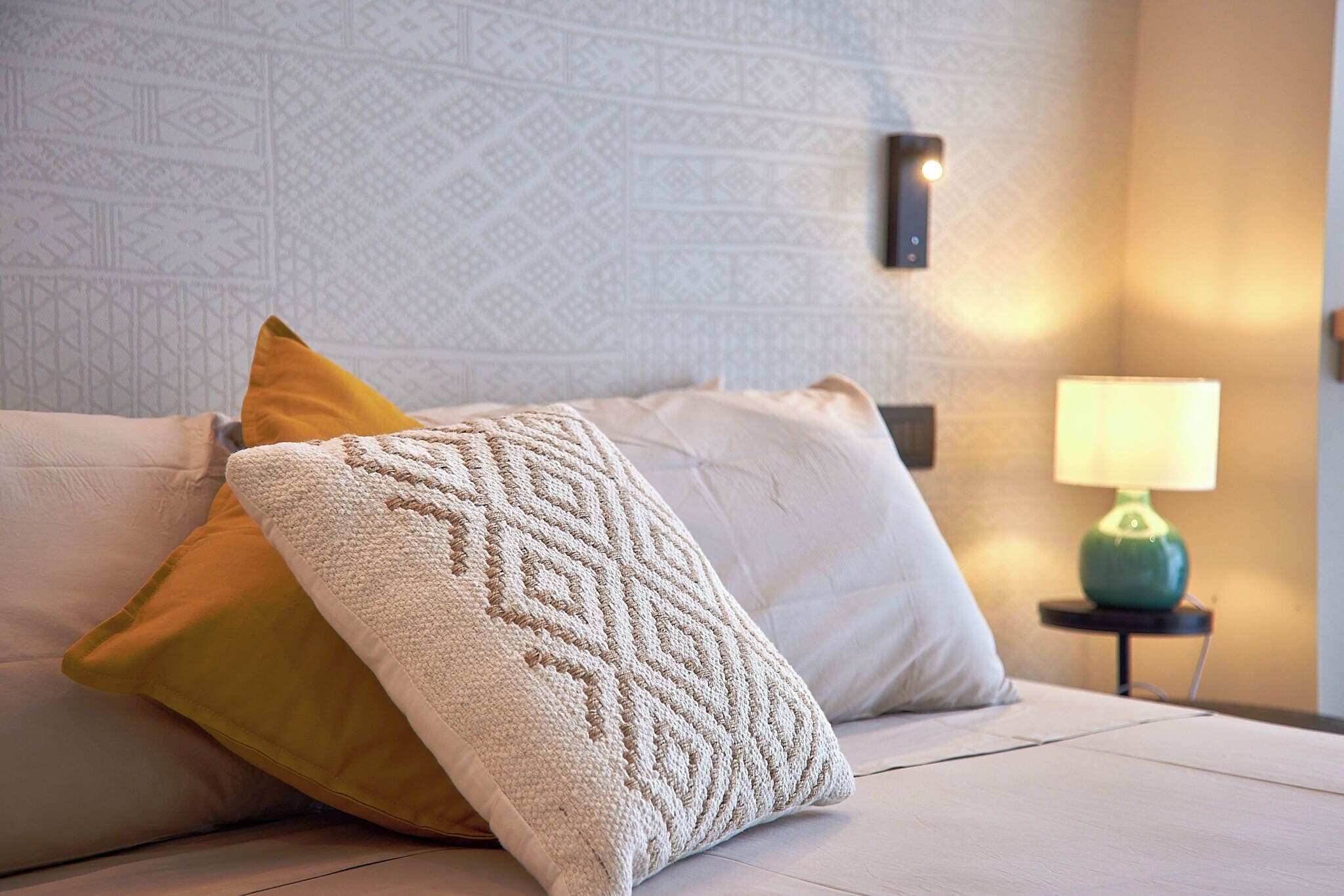 A neatly made bed with a patterned throw blanket and pillows, a wall-mounted light fixture, and a small table with a lamp.