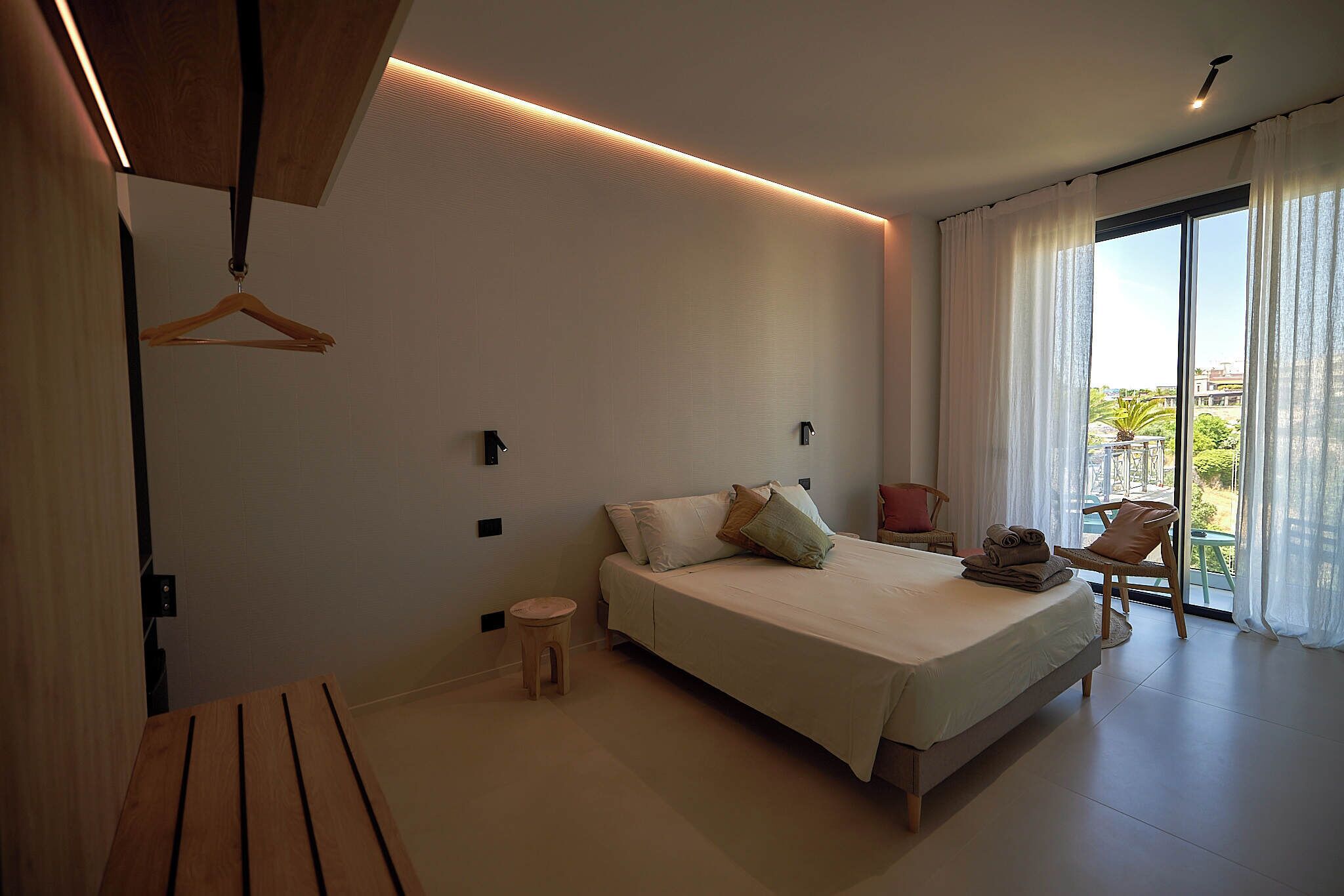 A modern bedroom with a large bed, a small stool, a wooden bench, and a chair by a sliding glass door.