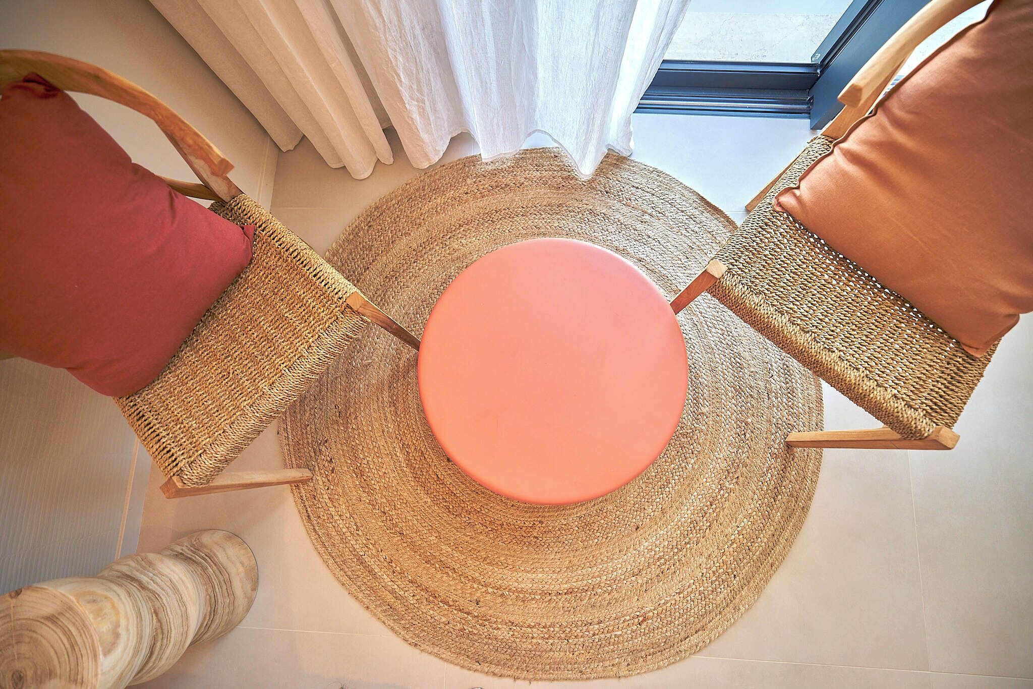 Une table ronde à la surface rose, entourée de deux chaises en osier avec des coussins marron, posée sur un tapis tissé.