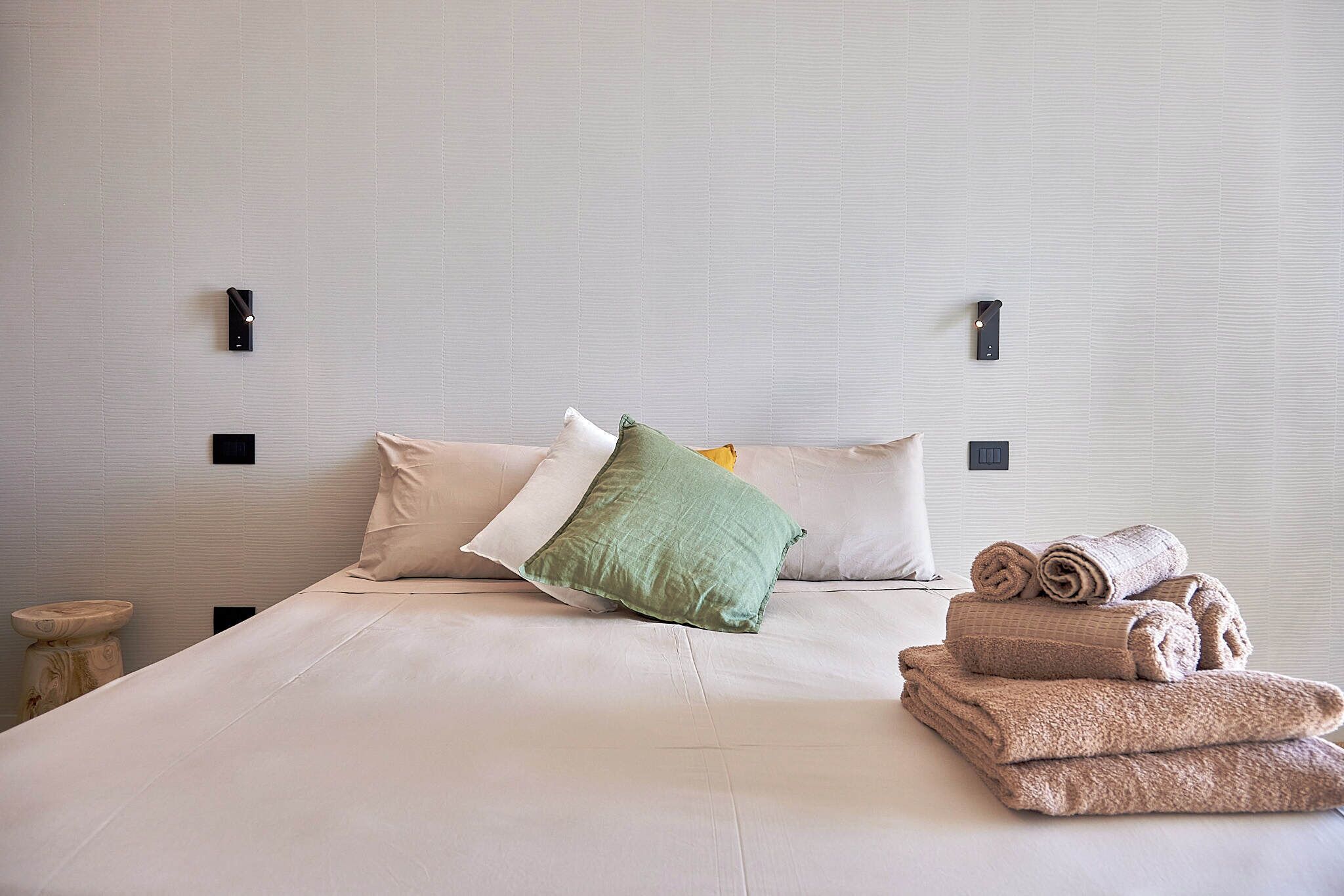 A neatly made bed with pillows and rolled towels, a bedside table, and wall-mounted light fixtures.