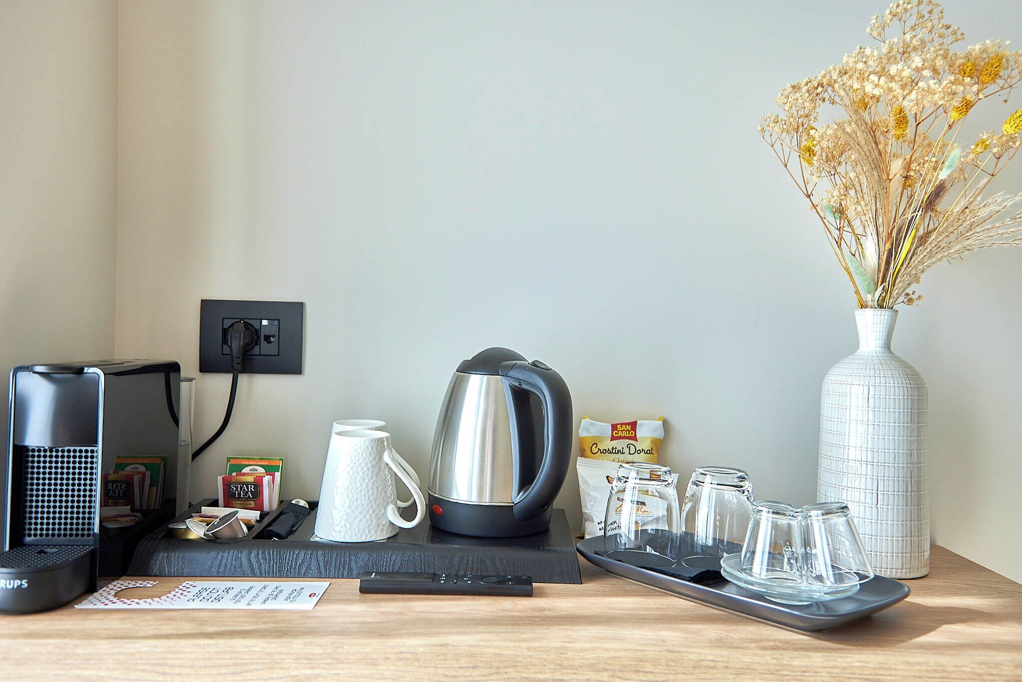 Une table de petit-déjeuner joliment dressée, avec une machine à café, une bouilloire, des tasses et un vase rempli de fleurs séchées.