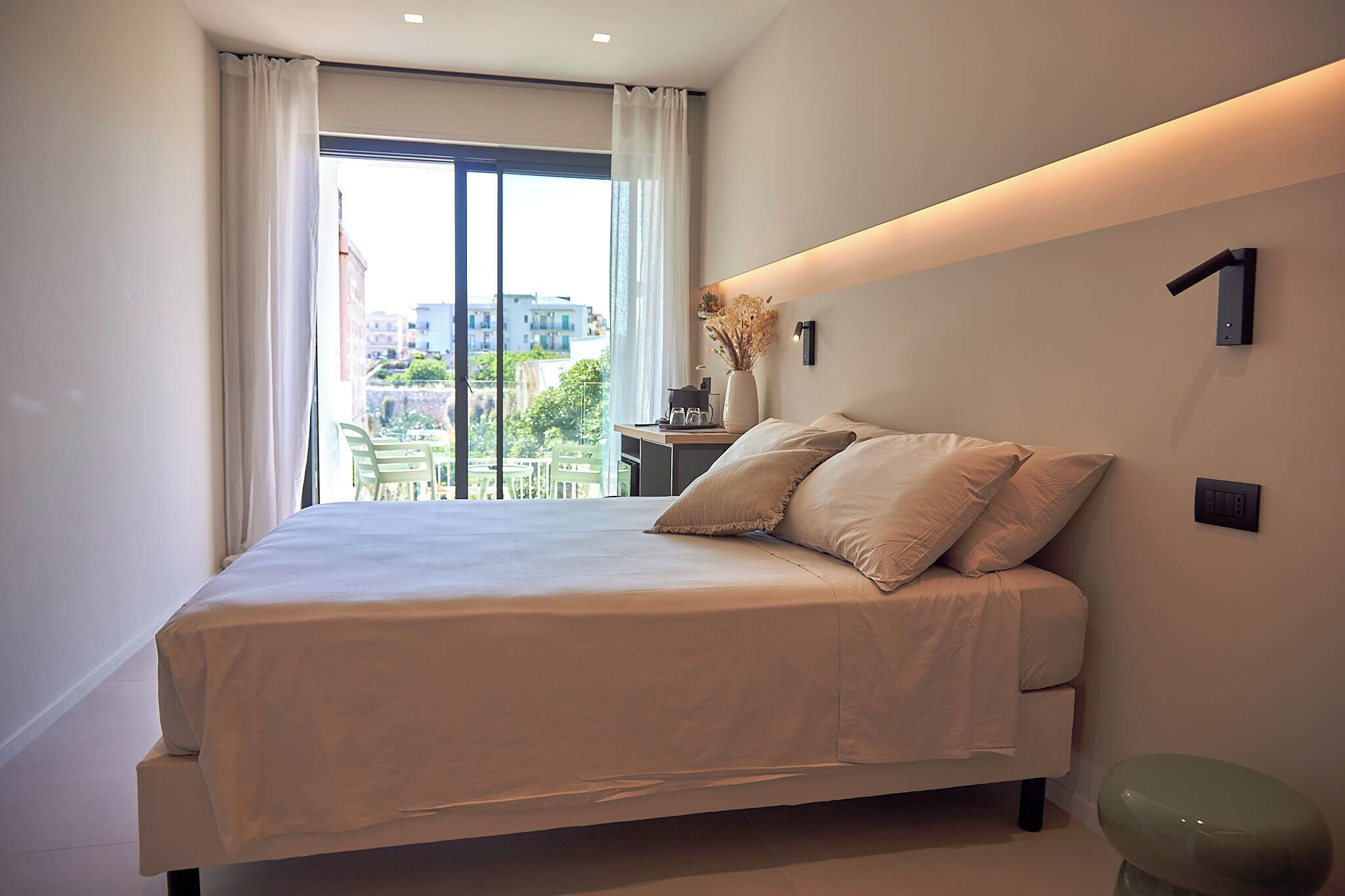 Un dormitorio moderno con una cama grande, una mesita de noche con un florero y vistas de la ciudad a través de un ventanal.