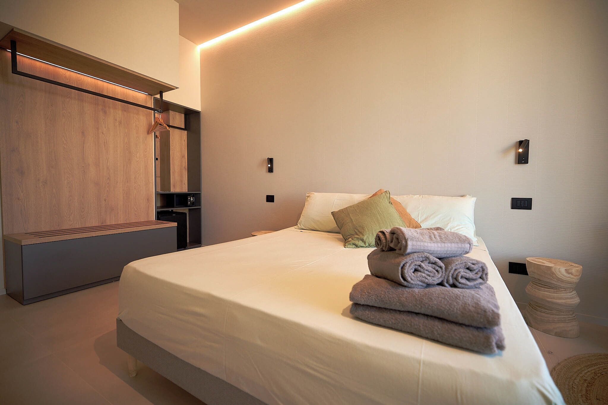 A modern hotel room with a large bed, a wooden wardrobe, and a small seating area.