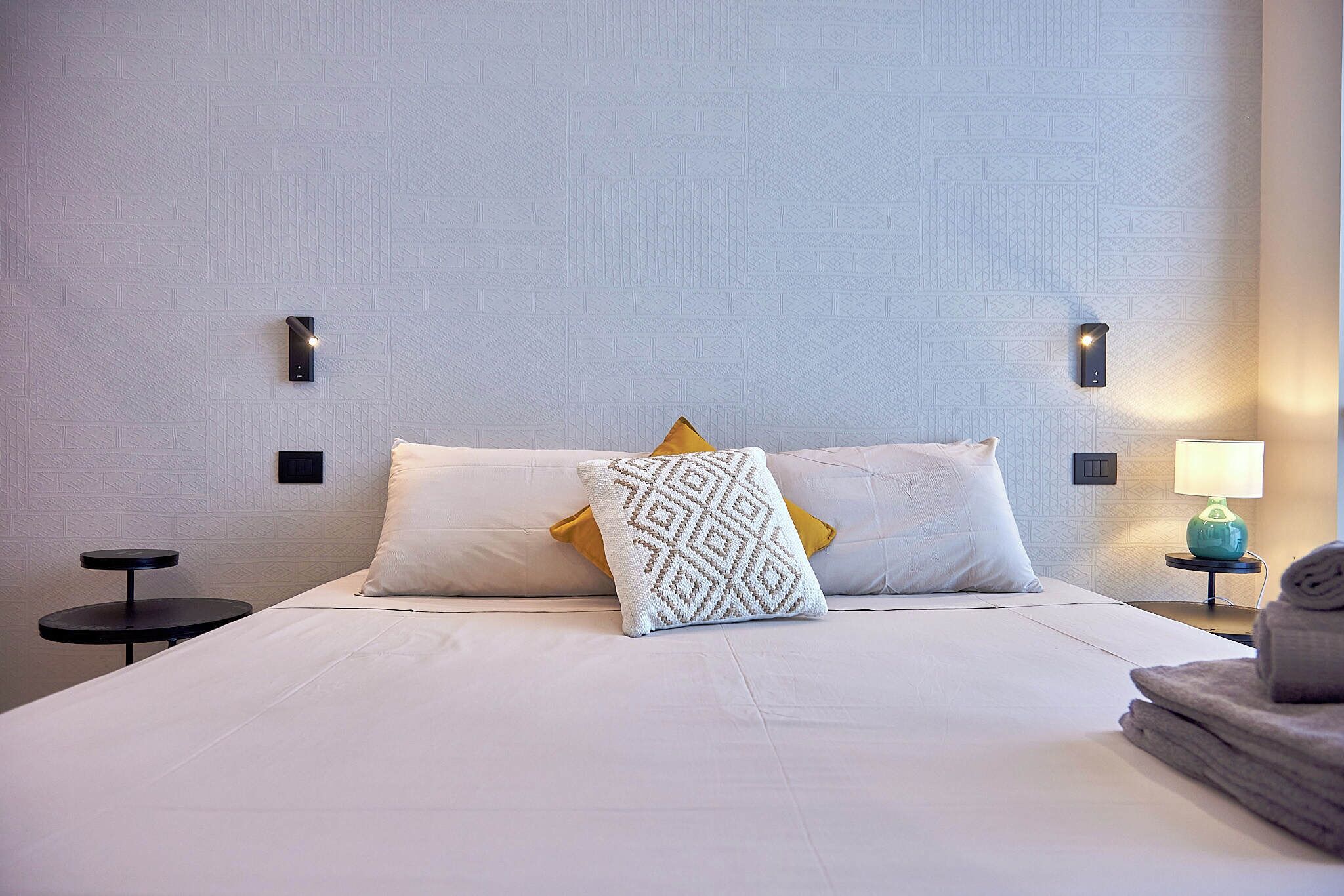 A neatly made bed with white linens, two pillows, a patterned throw, and a small bedside table with a lamp.