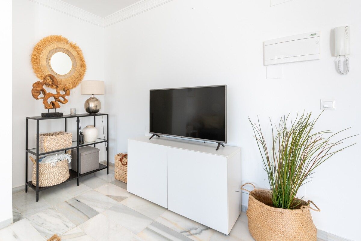 Een moderne woonkamer met een flatscreen televisie, een wit kastje, rieten manden, een ronde spiegel en een potplant.