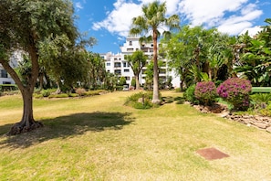 A well-maintained garden with various trees and shrubs, a clear sky, and a building with multiple balconies in the background. - Jardines Puerto Banús – Elegance and Comfort in Marbella’s Heart (Marbella)