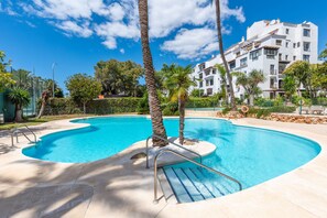 A swimming pool with a curved design, surrounded by palm trees and a building with balconies. - Jardines Puerto Banús – Elegance and Comfort in Marbella’s Heart (Marbella)