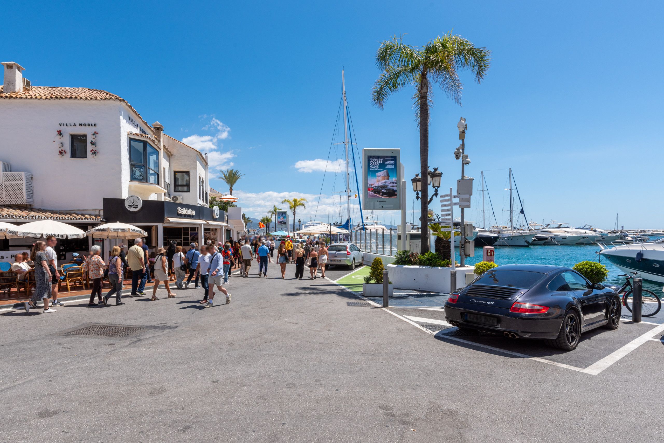 Un puerto deportivo con barcos, un coche negro y gente caminando por la calle.