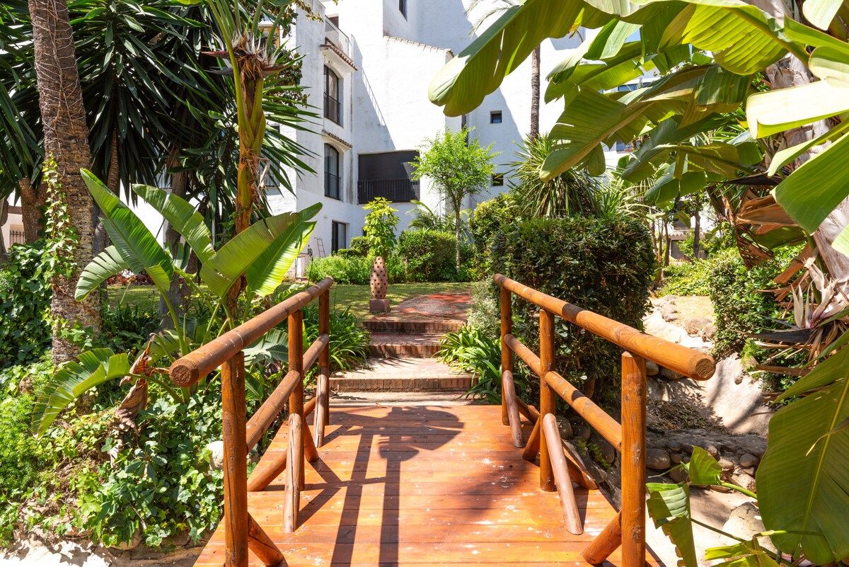A wooden pathway leading to a building with a white facade, surrounded by lush tropical vegetation.