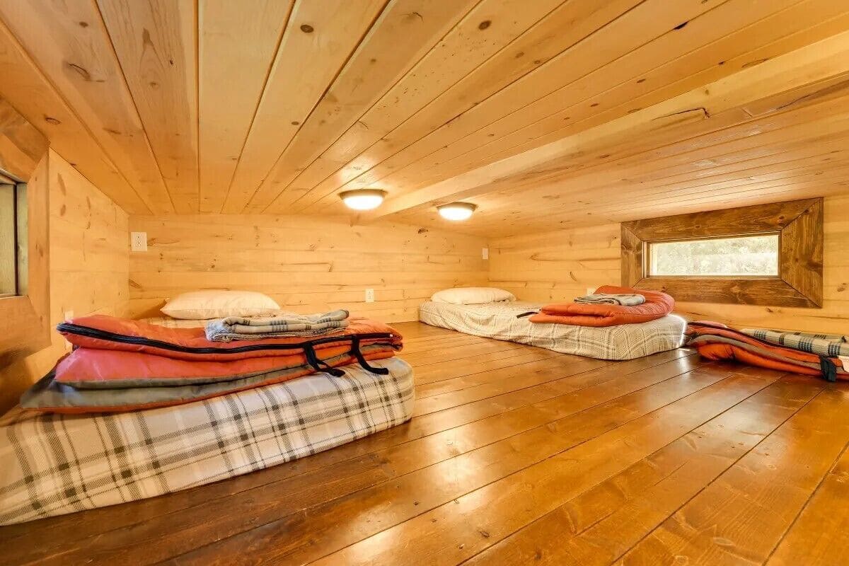 A cabin interior with wooden walls and floors, two beds with plaid bedding, and a window providing natural light.