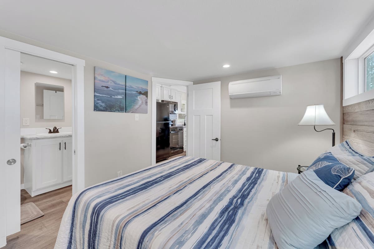 A bedroom with a bed, a nightstand, a lamp, a painting, and a view of the kitchen.