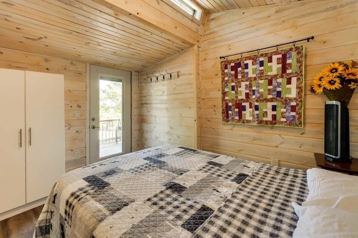 A cozy room with a wooden ceiling, a quilted bedspread, a quilt hanging on the wall, and a vase with sunflowers.