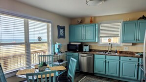 Cocina con gabinetes color turquesa, lavavajillas de acero inoxidable, microondas y cafetera. Hay una mesa de comedor con sillas y una alfombra en el suelo.