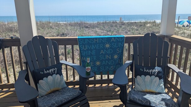 Zwei Adirondack-Stühle mit 'happy'-Kissen auf einer Holzterrasse mit Blick auf einen Strand.