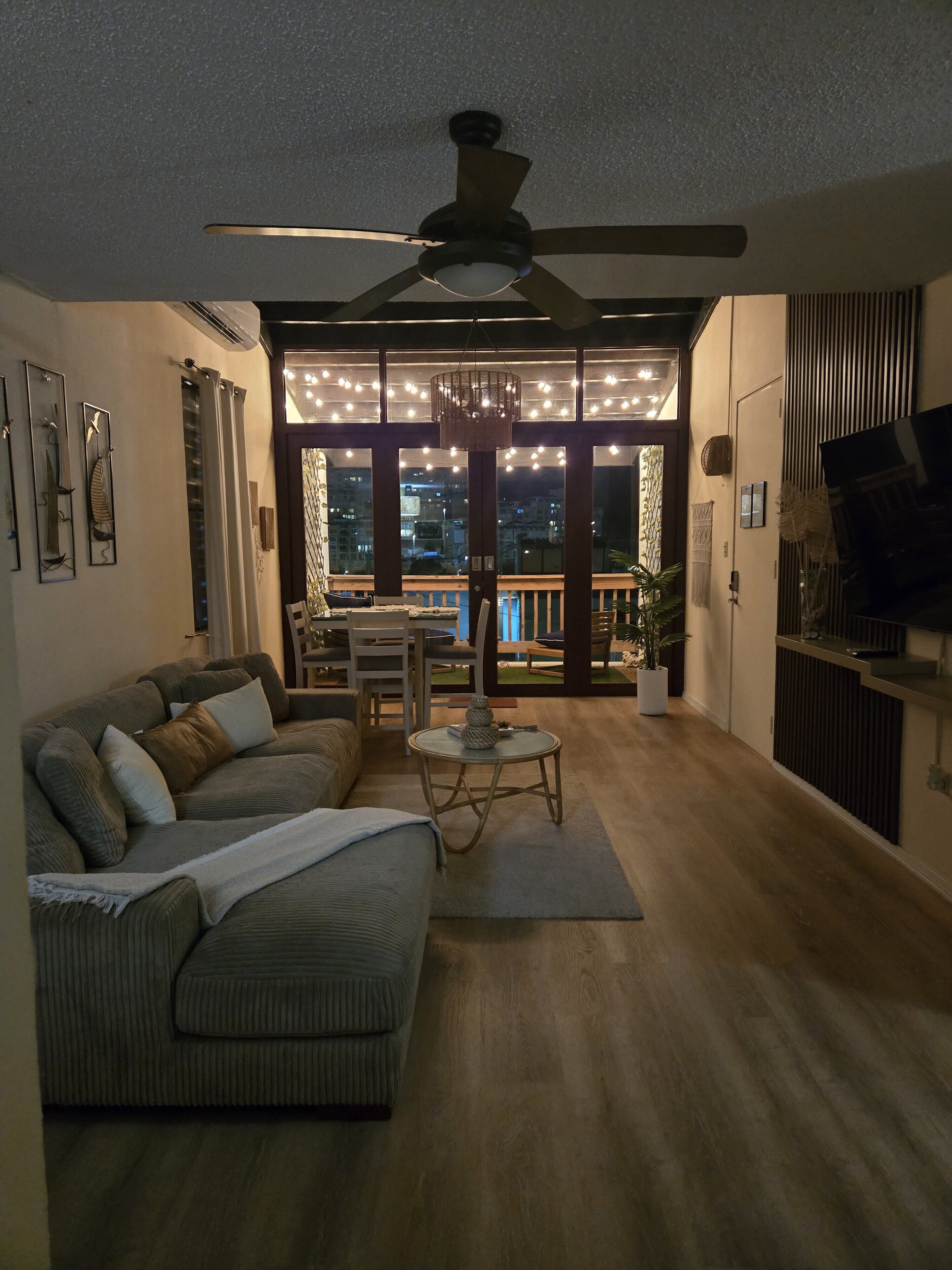A living room with a sectional sofa, a dining table with chairs, a ceiling fan, and a view of the city through large windows.