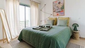 A bedroom with a green bedspread, a painting on the wall, and a small bedside table with a green lamp.