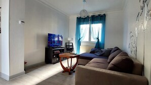 Un salon avec un canapé, un téléviseur, une petite table et une fenêtre avec des rideaux.
