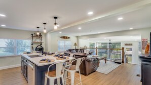 A spacious kitchen with a central island, a dining area with chairs, a living room with a sofa, and a view of a water body through large windows. - The Falling Pecan Lakehouse  (Granbury)
