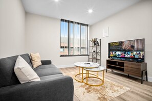 Ein modernes Wohnzimmer mit einem grauen Sofa, einem Couchtisch, einem Fernseher auf einem Holzständer und einem Fenster mit Jalousien.