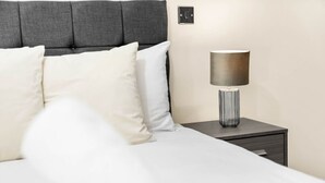 Ein ordentlich bezogenes Bett mit weißen Leinen und zwei Kissen, eine moderne Lampe auf einem Nachttisch und eine schlichte Wand mit Lichtschalter.
