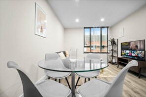 Ein modernes Wohnzimmer mit Glastisch und weißen Stühlen, ein großes Fenster mit Blick auf die Stadt, ein Fernseher auf einem Holzboden und ein gerahmtes Bild an der Wand.