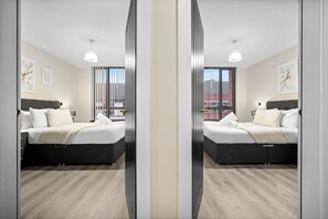 Ein modernes Schlafzimmer mit Spiegelblick, das ein Bett, Nachttische und ein Fenster mit Jalousien zeigt.