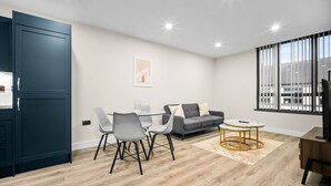 Ein modernes Wohnzimmer mit einem grauen Sofa, einem Glastisch mit Stühlen, einem Couchtisch und einem Fernseher auf einem Holzgestell.