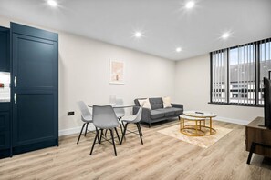 Ein modernes Wohnzimmer mit einem grauen Sofa, einem Glastisch mit Stühlen, einem Couchtisch und einem Fernseher auf einem Holzgestell.