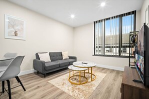 A modern living room with a grey sofa, two round tables with gold legs, a TV on a wooden stand, and a large window with blinds. - Luxury 2 Bed Apartment - Smart TV - WIFI -  Jewellery Quarter (Birmingham)