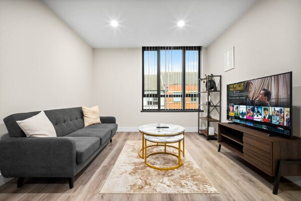 A modern living room with a grey sofa, a round coffee table, a TV on a wooden stand, and a large window with blinds.