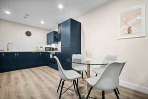 A modern kitchen with dark blue cabinets, a white tiled backsplash, and a round glass table with four white chairs.