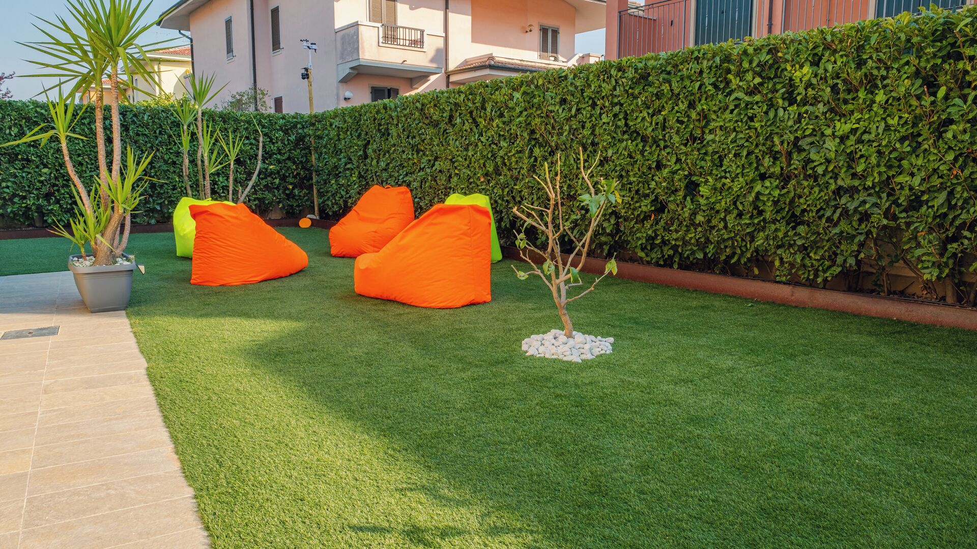 Un césped cuidado con tres sillones orejeros naranjas, un árbol pequeño y un seto.