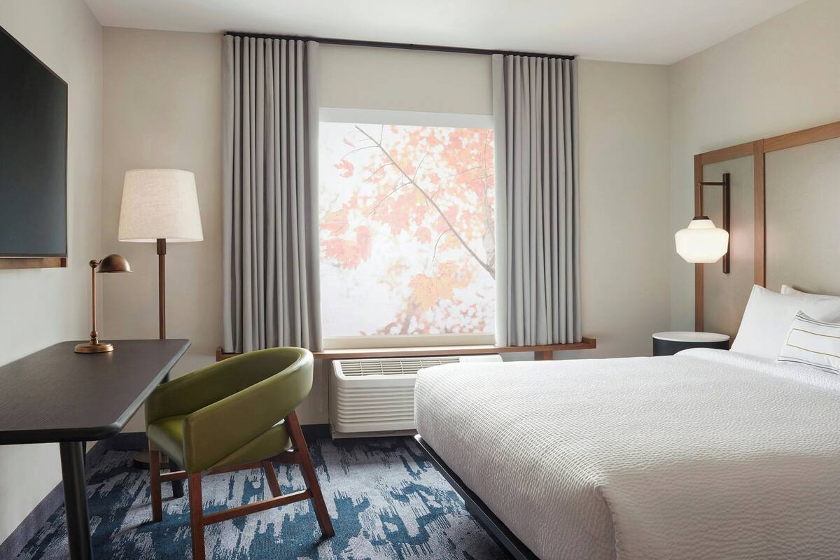 A hotel room with a bed, a desk, a chair, a lamp, a nightstand, and a window with curtains.