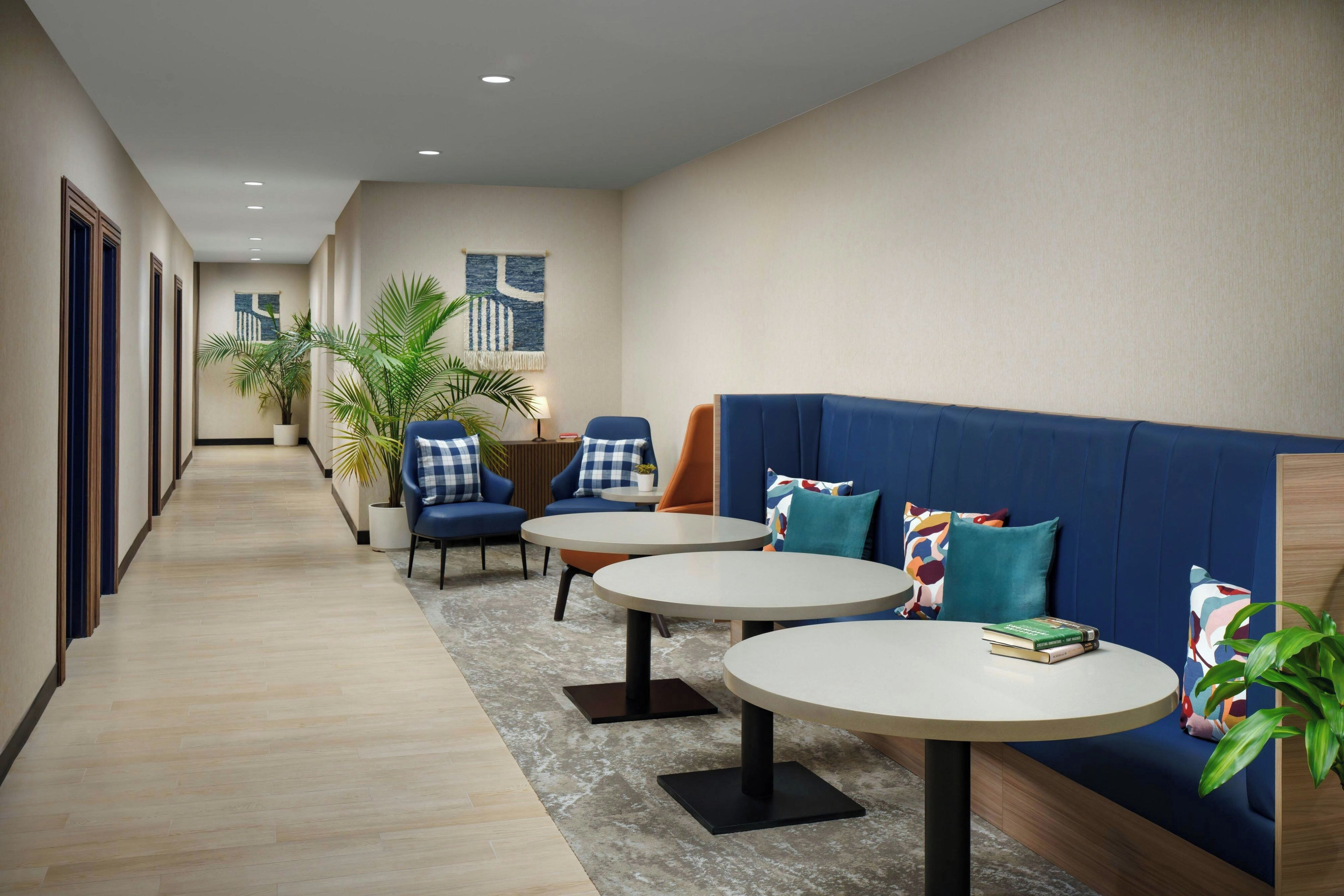 A hotel lobby with a long corridor, seating area with blue sofas and chairs, and a small table with books.